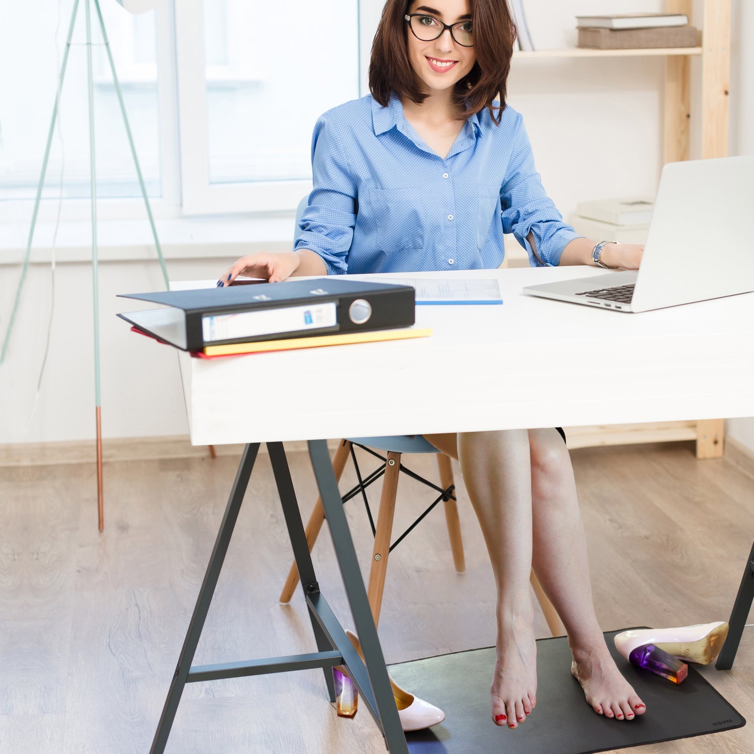 Eine Frau sitz im Büro am Schreibtisch und lächelt. Sie berührt mit den Füssen eine Erdungsmatte, die unter dem Schreibtisch liegt.